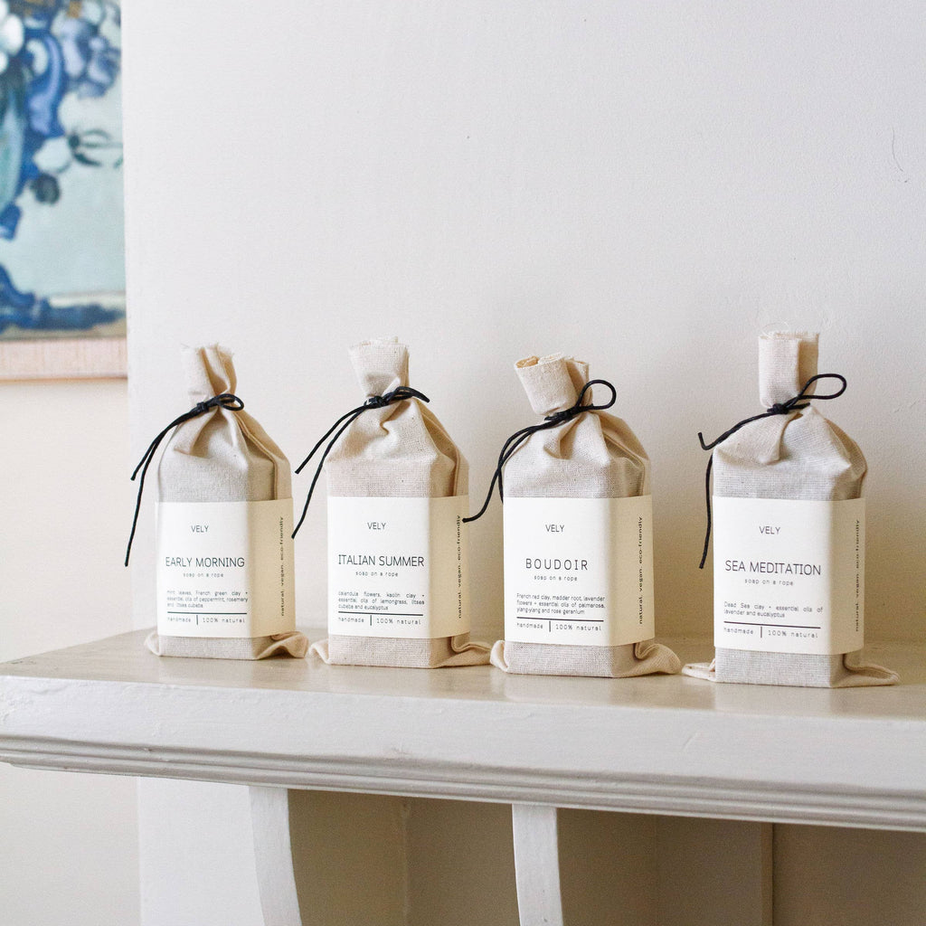 Four white cotton pouches with VELY soap bars with black ribbons on a marble surface against a light grey wall.