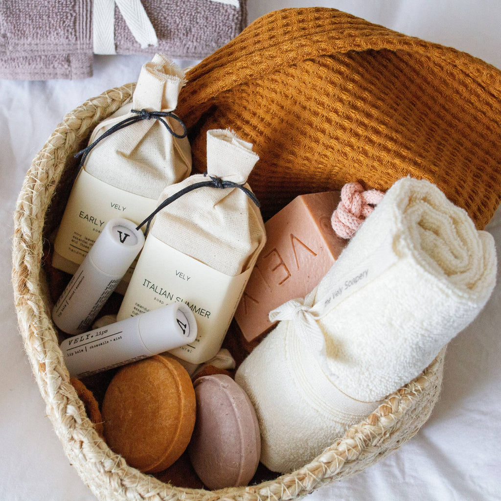 Basket with various VELY soap bars, lip balms and a towel on a white background