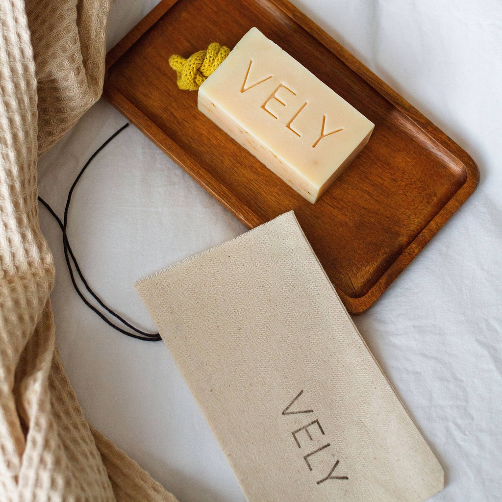 Bar of VELY soap on a wooden tray with a matching soap bag on a white background